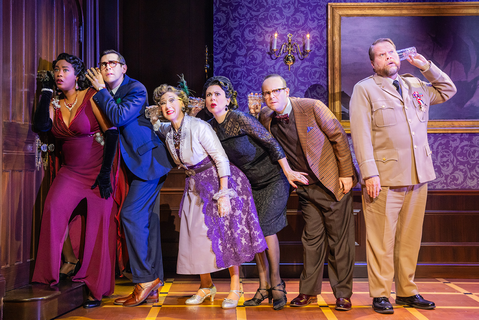 The Company of the North American tour of CLUE - photo by Evan Zimmerman for MurphyMade<br><br>Six actors stand on stage in a line on one side of a closed wooden door, five of them are crouched close together with their ears pressed against glasses, trying to hear a conversation inside the closed door. The other actor is standing facing away from the rest, his glass pressed to his own ear, but the other end is connected to nothing.