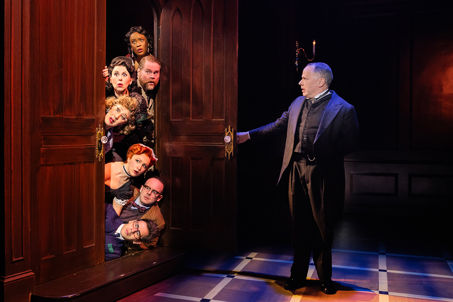 The Company of the North American tour of CLUE - photo by Evan Zimmerman for MurphyMade<br><br>Seven actors lay stacked on top of each other horizontally, peeking out from behind an open door. Only their surprised faces are visible outside of the door frame. Across from them, a man in a suit has one arm outstretched, having just opened the door.