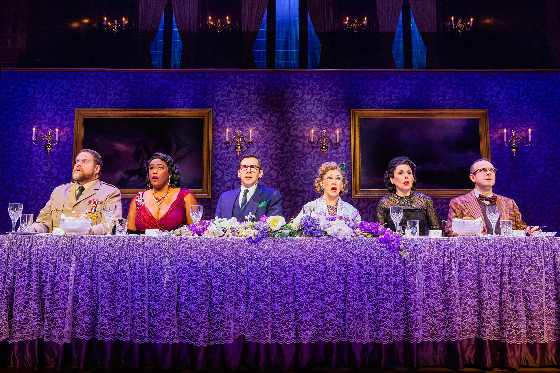 The Company of the North American tour of CLUE - photo by Evan Zimmerman for MurphyMade<br><br>Six actors in formal attire sit all on the same side behind a long fancy dinner table. Each actor is staring forward into the distance with a shocked and perplexed look.
