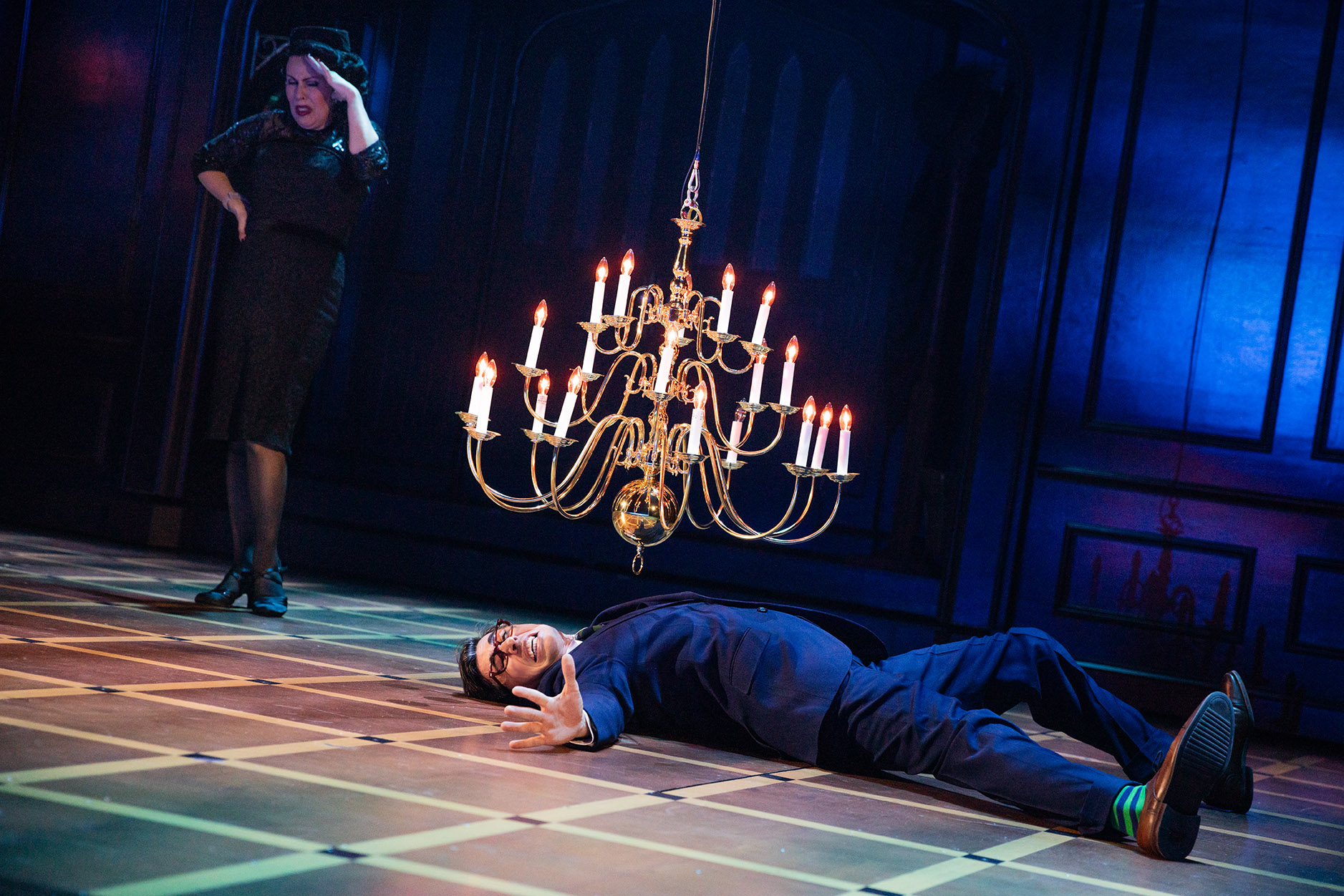 John Shartzer and Tari Kelly in the North American tour of CLUE - photo by Evan Zimmerman for MurphyMade<br><br>A man is lying spread out on the floor, mouth agape as a falling chandelier hangs over him. A woman in all black stands in the foreground with her hand covering her eyes. 